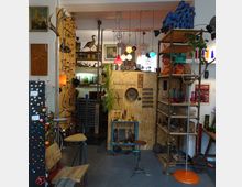 Innenansicht eines Antiquitäten- und Vintage-Ladens mit Regalen, die verschiedene Objekte wie Glasgefäße, Dekorationsgegenstände und Lampen zeigen. Die Wände sind mit Holzplatten verkleidet, es hängen bunte Pendelleuchten von der Decke, und es gibt verschiedene Möbelstücke sowie auffällige Dekorelemente wie ein Geweih und alte Porträts.