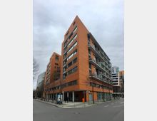 Moderner, mehrstöckiger Wohn- und Bürokomplex mit orangefarbener Fassadenverkleidung, großen Fensterbändern und durchgehenden Balkonen an der Straßenecke. Im Vordergrund verlaufen breite Gehwege mit kahlen Straßenbäumen, Fahrradständern und Straßenbeschilderung an einer Kreuzung.