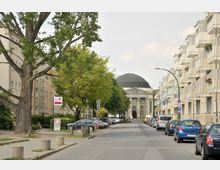 Straßenansicht mit geparkten Autos und Wohngebäuden auf beiden Seiten; im Hintergrund ein neoklassizistisches Gebäude mit einer großen Kuppel, umgeben von Bäumen.