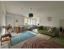 Wohnzimmer mit heller, moderner Einrichtung. Ein grünes Sofa, bunte Teppiche und ein Schreibtischbereich mit großen Fenstern dominieren den Raum, der durch Dekorationen wie Blumen, Bilderrahmen und eine Stehlampe ergänzt wird.