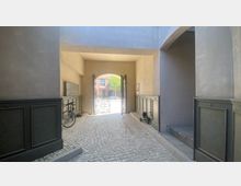 Gepflasterter Durchgang mit hohen, hellen Wänden und grün lackiertem Wandpaneel. Ein offenes, kunstvoll verziertes Eisentor führt zu einem sonnigen Hof mit Baum und Backsteingebäude im Hintergrund; ein Fahrrad und ein Kinderdreirad stehen seitlich.