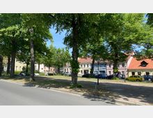 Eine von Bäumen gesäumte Straße mit einer kleinen Grünfläche, auf der Bänke und Spaziergänger zu sehen sind. Im Hintergrund stehen mehrfarbige, zweigeschossige Wohn- und Geschäftshäuser mit Satteldächern.