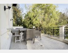 Terrasse mit einem Holztisch und geflochtenen Sitzmöbeln, umgeben von einem dekorativen Metallgeländer; im Hintergrund dichter Baumbestand mit grünem Laub.