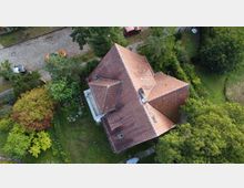 Luftaufnahme eines Hauses mit rotem Ziegeldach und mehreren Dachflächen, umgeben von dichtem Grün und Gartenflächen. Am oberen Bildrand verläuft eine Kopfsteinpflasterstraße mit parkenden Autos entlang eines Gehwegs.