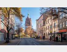 Straßenszene mit Bäumen und mehrstöckigen Gebäuden auf beiden Seiten, im Hintergrund ragt der Turm einer Kirche mit spitzem Dach und grünem Turmhelm hervor. Rechts ist eine Bushaltestelle mit wartenden Personen sichtbar, links ein moderner Backsteinbau.