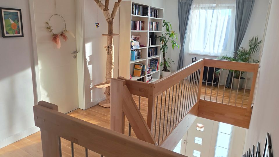 Heller Flur im Obergeschoss mit Holzparkett, Treppenöffnung und Holzgeländer mit Metallstäben, der Blick führt nach unten ins Treppenhaus. An den weißen Wänden stehen ein Bücherregal und Pflanzen, am Ende befindet sich ein großes Fenster mit Vorhängen.
