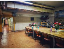 Innenraum eines schlichten Aufenthalts- oder Speiseraums mit langen Holztischen und Stühlen, auf den Tischen stehen Vasen mit Blumen und Thermoskannen. An der Decke verlaufen sichtbare Lüftungskanäle und Leuchtstoffröhren; rechts hängt ein großes Landschaftsbild, links führt eine offene Tür in einen langen Flur.