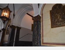 Treppenhaus in einem historischen Gebäude mit verzierten Marmorsäulen, weißen Rundbögen und einer dekorativen Wandplakette aus Metall. Eine große, antike Hängelaterne beleuchtet den Bereich.