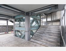 Treppenbereich in einem modernen, überdachten Gebäude mit Glas- und Stahlkonstruktion. Die Treppe aus grauem Stein führt neben verglasten Wänden mit Metallsäulen hinauf, umgeben von Geländern und einer klaren, industriellen Ästhetik.