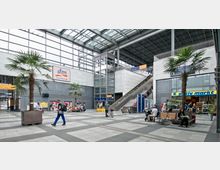 Helle Bahnhofshalle mit moderner Glas-Stahl-Konstruktion, Palmen in Pflanzkübeln und Geschäftseingängen, darunter ein Drogeriemarkt und ein Supermarkt. Eine breite Treppe und ein Fahrstuhl führen zu einer oberen Ebene.