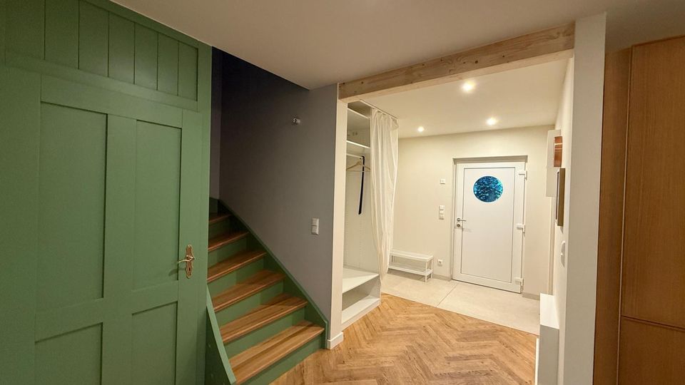 Eingangsbereich eines Hauses mit Holztreppe links, grüner Wandverkleidung und Fischgrätparkettboden. Rechts befindet sich eine weiße Haustür mit rundem Fenster und ein offener weißer Einbauschrank mit Vorhang.