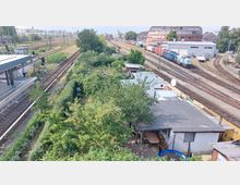 Blick auf einen Bahnhofsbereich mit Gleisanlagen, einer Bahnhofsplattform links und einem angrenzenden Schrebergarten mit dichtem Grün und kleinen Gartenhäusern. Im Hintergrund sind Güterzüge und ein Industriegebäude zu sehen.