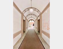 Ein schmaler, überdachter Durchgang mit gewölbter Decke und cremefarbenen sowie beigen Verzierungen, der zu einem offenen Hof mit Backsteingebäude im Hintergrund führt. Eine runde Lampe hängt zentral von der Decke, und an der rechten Wand ist orangefarbene Graffiti erkennbar.