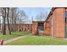 Backsteingebäude in einem parkähnlichen Umfeld mit großen Fenstern und mehreren Bäumen im Vordergrund. Im Hintergrund sind weitere Gebäude aus rotem Backstein mit großen Fenstern sichtbar.