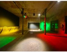 Ein leerer, industriell anmutender Raum mit grob verputzten Wänden und sichtbaren Backsteinen. Der Raum wird in gelbes, rotes und grünes Licht getaucht, mit einzelnen Säulen und einer schwarzen Tafelart an der Wand.