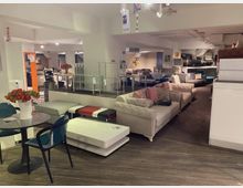 Innenraum eines Möbelgeschäfts mit mehreren Sofas, Sesseln und Tischen in unterschiedlichen Designs und Farben. Der Laden hat helle Wände, eine Holzoptik am Boden und moderne Beleuchtung; eine Glasbrüstung trennt verschiedene Ausstellungsbereiche.