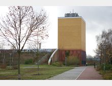 Ein großer rechteckiger Turm mit gelb-beiger Fassade und technischen Installationen auf dem Dach, umgeben von Bäumen und Grünflächen. Im Vordergrund führen Fuß- und Radwege entlang, und links ist eine Treppe zu sehen, die eine Böschung hinaufführt.