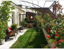 Dachterrasse mit begrünter Rasenfläche, seitlichen Pflanzkübeln mit roten und weißen Blumen, und einer Sitzgruppe aus Korbmöbeln im Hintergrund unter einer offenen Metallkonstruktion. Im Hintergrund ist ein Gebäude mit Ziegeldach sichtbar.