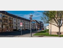 Straßenszene in einer Wohngegend mit mehrstöckigen, hell verputzten Wohnhäusern und mehreren geparkten Autos. Im Vordergrund ein Straßenschild mit Geschwindigkeitsbegrenzung auf 30 km/h und ein Baum auf einer Grünfläche neben einem Gehweg.