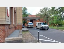 Backstein-Gebäude mit einer Einfahrt für Krankenwagen, die von einer Überdachung überspannt ist. Im Vordergrund sind zwei parkende Krankenwagen zu sehen, im Hintergrund befindet sich eine Grünfläche mit Bäumen.