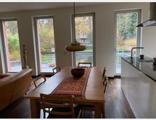 Offener Ess- und Küchenbereich mit großem Holztisch, Stühlen und einer Hängelampe; rechts eine moderne Küchenzeile mit Spüle. Mehrere bodentiefe Fenster und eine Glastür geben den Blick auf eine Terrasse und einen Garten mit Pflanzen frei.