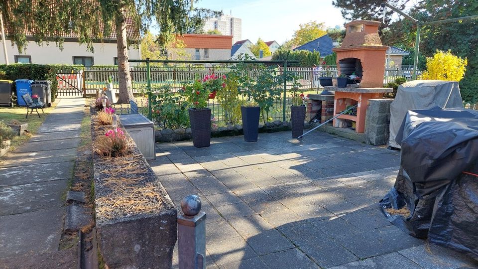 Gepflasterte Garten- und Terrassenfläche mit Pflanzkübeln und Blumenbeet entlang eines Gehwegs, eingefasst von niedrigen Steinmauern und einem Metallzaun. Rechts steht ein gemauerter Grillkamin neben abgedeckten Gartenmöbeln; im Hintergrund sind Wohnhäuser und Bäume zu sehen.