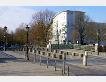 Straßenszene mit gepflastertem Gehweg, dekorativen Straßenlampen und runden Betonpollern. Im Hintergrund steht ein weißes mehrstöckiges Gebäude neben einer grünen Brücke, umgeben von laubfreien Bäumen.