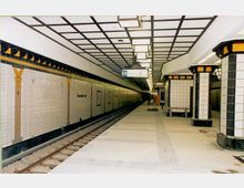 U-Bahn-Station mit hell gefliesten Wänden und gelben Dekorelementen, sowie schwarz-weißen, verzierten Säulen. Der Bahnsteig ist leer, und die Schienen verlaufen parallel zur Wand, während von der Decke rechteckige Lampen hängen.