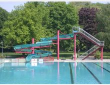 Ein Freibad mit einer großen Wasserrutsche aus grünem Kunststoff, gestützt von roten Metallpfeilern und zugänglich über eine Treppe. Im Hintergrund sind grüne Bäume und eine parkähnliche Landschaft zu sehen.