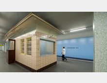 U-Bahnhof mit rechteckigem Kiosk, der mit beigefarbenen Kacheln verkleidet ist; daneben eine blaue Wand mit der Aufschrift "Afrikanische Str." und eine Person, die daran vorbeigeht. Deckenbeleuchtung und grauer Boden prägen die Station.