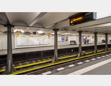 U-Bahnhof mit der Aufschrift „Theodor-Heuss-Platz“ an einer hell beleuchteten Bahnsteigwand. Der Bahnsteig ist von rechteckigen Säulen getragen, die Gleise sind von gelben Sicherheitsstreifen umsäumt, und im Hintergrund sind Sitzbänke, Informationsschilder und Passagiere zu sehen.