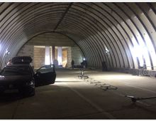 Innenaufnahme eines großen, bogenförmigen Hangars mit Betonwänden und -boden. Im Vordergrund steht ein Auto mit geöffneter Tür, während im Hintergrund Scheinwerfer und Filmtechnik sichtbar sind. Zwei helle Banner hängen an der Wand im hinteren Bereich.