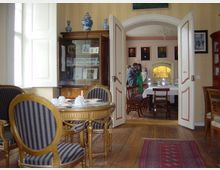 Ein historischer Raum mit Holzboden, in dem ein eleganter runder Tisch mit goldenen Stühlen im Vordergrund steht. Durch geöffnete Doppeltüren ist ein weiterer Raum mit weiß gedecktem Tisch, Porzellanausstellungsstücken und gerahmten Porträts an den Wänden sichtbar.