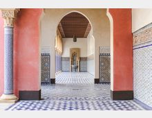 Innenansicht eines marokkanischen Gebäudes mit einem überdachten Gang, dekorativen Bögen und reich verzierten Fliesen in blau-weißen geometrischen Mustern. Am Ende des Ganges befindet sich ein kleiner Brunnen vor einer aufwendig verzierten Nische.