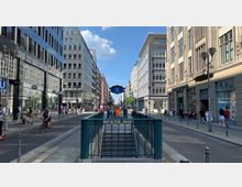 Straßenansicht in einer Stadt mit modernen Gebäuden auf beiden Seiten, einem Gehweg und einer Fahrbahn. Im Vordergrund befindet sich ein Eingang zur U-Bahn-Station "Stadtmitte" mit einem blauen Schild, und Menschen gehen oder fahren Fahrrad entlang der Straße.