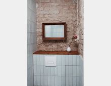 Ein kleiner Badezimmerbereich mit einer Wand aus unbehandeltem Ziegelstein und hellblauen Fliesen. Darüber ein Holzregal mit einer weißen Vase und einer vertrockneten Blume sowie ein rechteckiger Spiegel in einem rustikalen Holzrahmen.