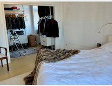 Schlafzimmer mit einem weißen Bett, bedeckt mit einer Felloptik-Decke. Im Hintergrund ein begehbarer Kleiderschrank mit einer Kleiderstange, Regalen, einer Leiter und einem geflochtenen Wäschekorb; helle Wände und Holzboden prägen den Raum.