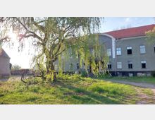 Großes, zweistöckiges Gebäude mit grauer Fassade und rot gedecktem Dach, zahlreiche Fenster, einige davon beschädigt oder geöffnet. Im Vordergrund ein großer Baum mit hängenden Ästen sowie eine Wiese und ein gepflasterter Weg, der in Richtung des Gebäudes führt.