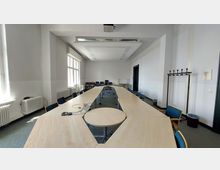 Großer, heller Konferenzraum mit weißen Wänden, langen Tischen in U-Form und blauen, gepolsterte Stühlen. Im Raum befinden sich eine große Fensterfront auf der linken Seite, eine Garderobe rechts und ein Beamer an der Decke.