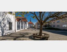Eine gepflasterte Platzfläche mit einem zentral stehenden Baum, der von einer runden Holzbank umgeben ist. Im Hintergrund sind weiße und beige Gebäude mit roten Ziegeldächern sowie ein Kirchturm mit schwarzer Spitze zu sehen.