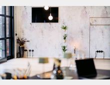 Minimalistisch eingerichteter Innenraum mit einer Betonwand, an der ein schwarzer Bildschirm und zwei Hängelampen angebracht sind. Links ein großes Fenster mit schwarzem Rahmen, rechts ein Pflanzenständer mit mehreren kleinen Pflanzen, daneben eine brennende Kerze auf einem kleinen Tisch.