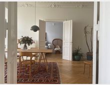 Ein heller Raum mit Holzboden und Stuckdecke, eingerichtet mit einem Holztisch, umgeben von Stühlen, auf einem großen, gemusterten Teppich. Im Hintergrund sind geöffnete Doppeltüren, ein weiterer Raum mit einem Korbsessel und Pflanzen sichtbar.