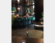 Elegante Bar mit einer dunklen, modernen Einrichtung. Eine geschwungene Bartheke mit grünen Polsterhockern wird von Stehlampen mit grünen Lampenschirmen beleuchtet, umgeben von schwarzem Interieur und floralen Wandverkleidungen.