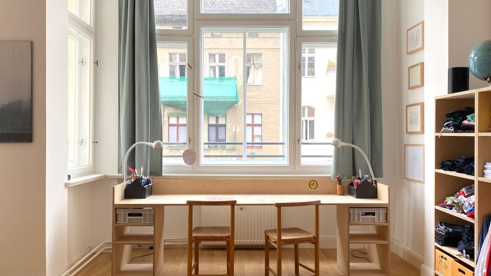 Heller Arbeitsbereich mit langem Holztisch und zwei Stühlen vor großen Sprossenfenstern mit Vorhängen, ausgestattet mit Schreibtischlampen und Stifteboxen. Rechts steht ein offenes Regalsystem mit Fächern, links sind weitere Fenster und ein Wandbild zu sehen.