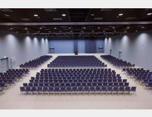 Großer Konferenzsaal mit mehreren Reihen ausgerichteter blauer Stühle, einer Bühne mit einem Rednerpult im Hintergrund und hellgrauen Wänden. Die Decke ist mit schwarzen Paneelen und eingebauten Leuchten ausgestattet.