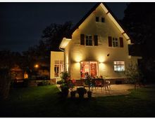Ein beleuchtetes Einfamilienhaus bei Nacht mit hellen Wänden und braunen Fensterläden. Im Vordergrund befindet sich eine Terrasse mit Stühlen, Pflanzen und einem angrenzenden gepflegten Rasen, umgeben von Bäumen im Hintergrund.