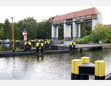 Schleusenanlage mit einem historischen Hebewerkgebäude, flankiert von gelb-schwarz markierten Pollern im Wasser. Im Hintergrund sind grüne Bäume und ein rotes Ziegeldach des Gebäudes zu sehen.