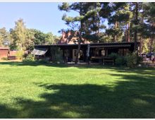 Ein schwarzes, einstöckiges Holzhaus mit vorgelagerter Terrasse und Markise steht inmitten eines Gartens mit Rasenfläche, umgeben von hohen Kiefern und Sträuchern. Im Hintergrund sind weitere Häuser und kleine Holzbauten zu sehen.