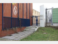 Backsteinfassade mit blauen Metallgittern und einer Treppe, die zu einem Metalltor führt. Rechts befinden sich eine grün verkleidete Wand und weiße Pfeiler mit Graffiti, umgeben von einer kleinen Rasenfläche.