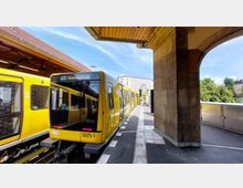 Eine gelbe U-Bahn steht an einem teilweise überdachten Bahnsteig einer erhöhten Station. Im Hintergrund sind ein historisches Gebäude und grüne Bäume unter einem blauen Himmel sichtbar.